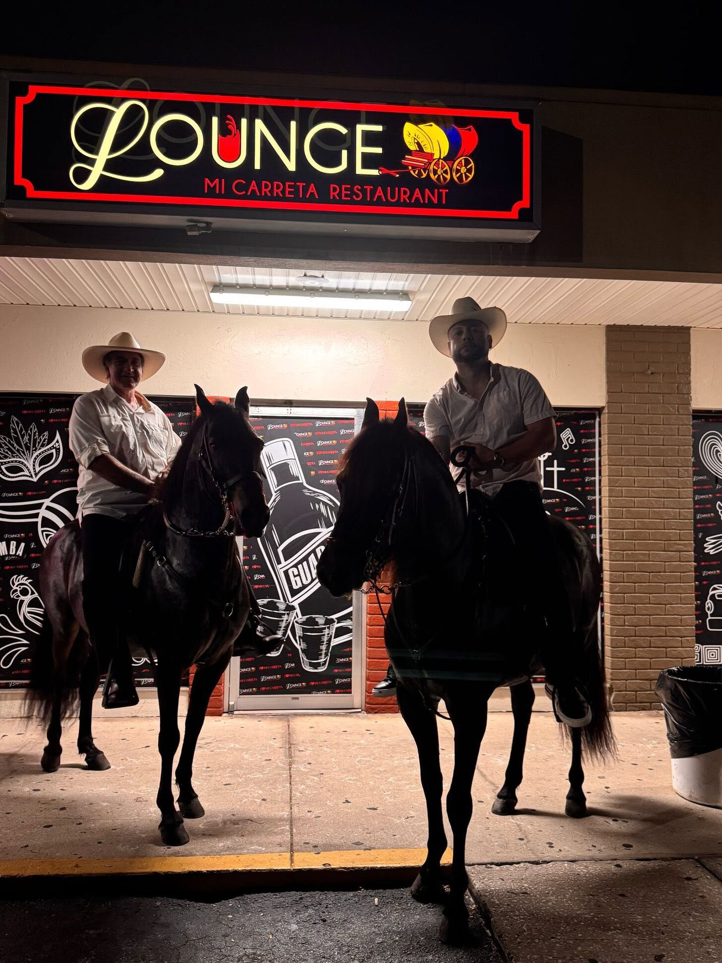 Two riders on horseback under the Lounge sign at night