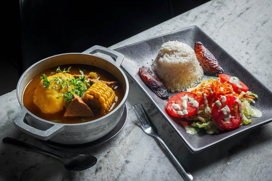 A pot of soup with corn beside a plate of rice, plantain and salad