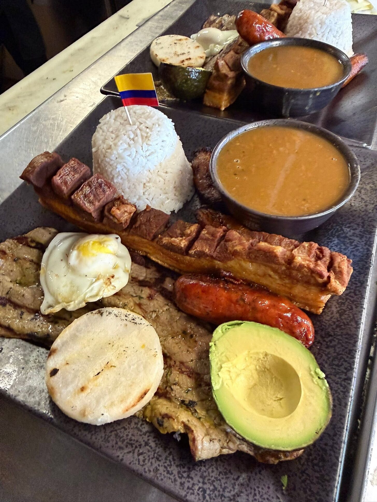 A loaded bandeja on a slate board with chicharrón, chorizo, egg, avocado, arepa and rice