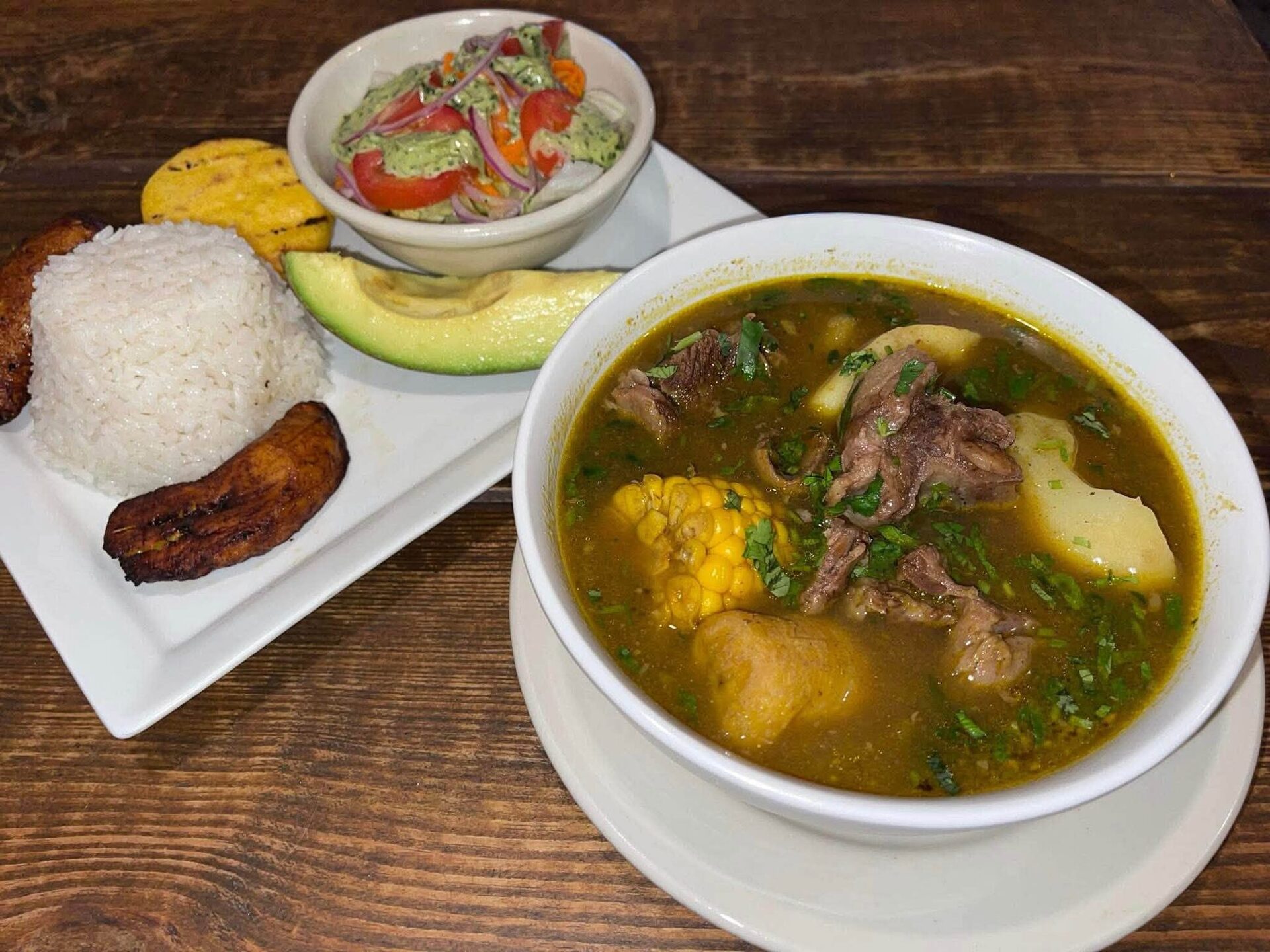 A bowl of Colombian soup with rice, avocado, a plantain and a small salad on a wooden table